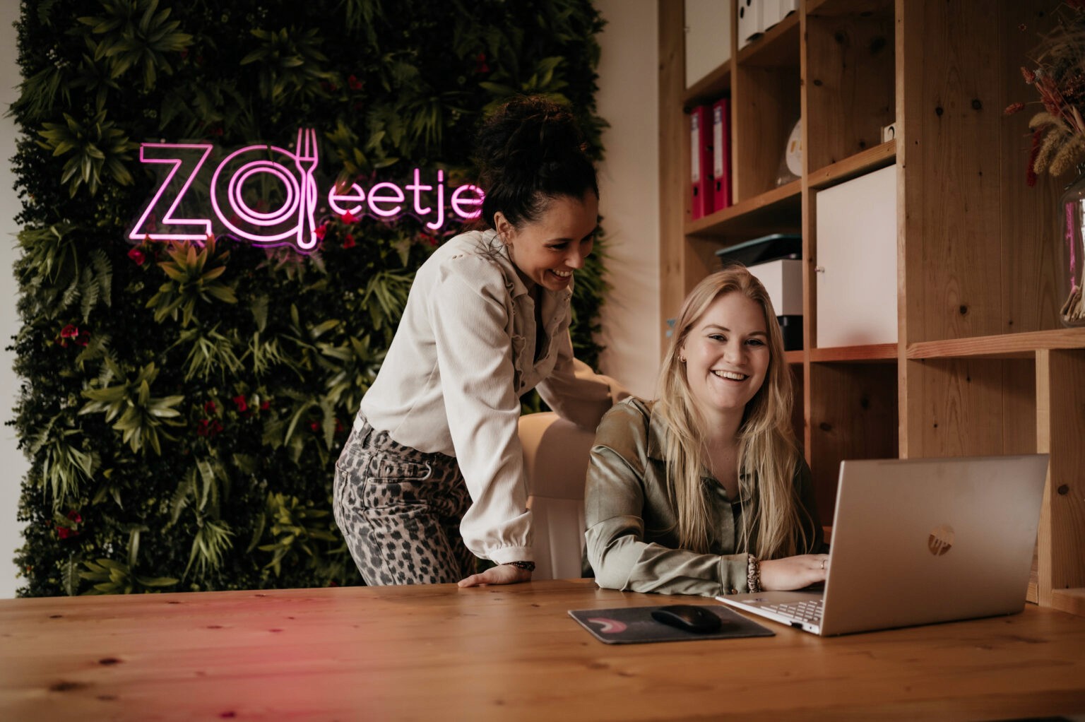 A person stands behind a woman at a desk, both engaged with a laptop in a stylish office space.