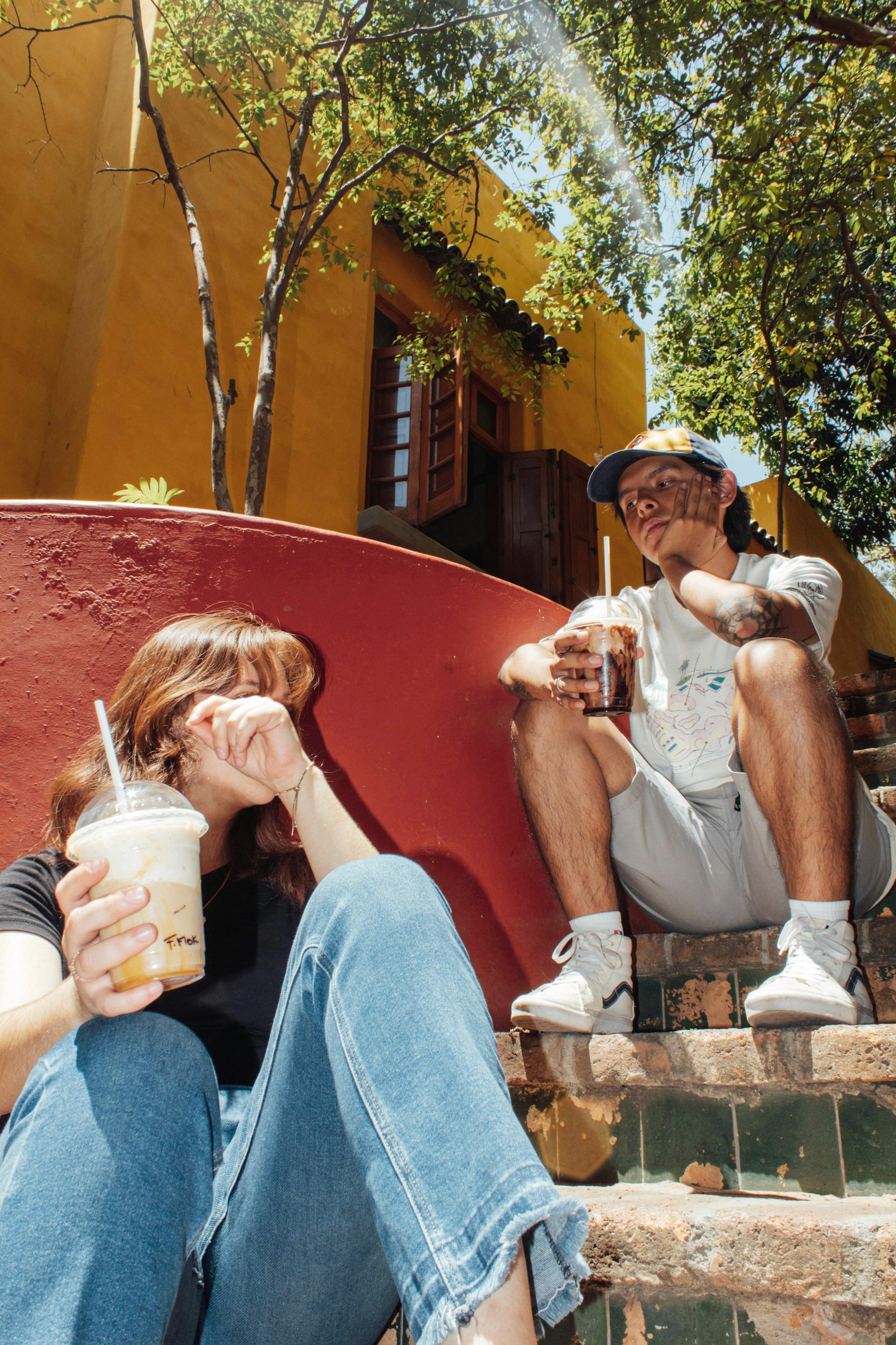 Two young adults sitting on outdoor brick steps in the sun, both holding large iced coffee drinks.