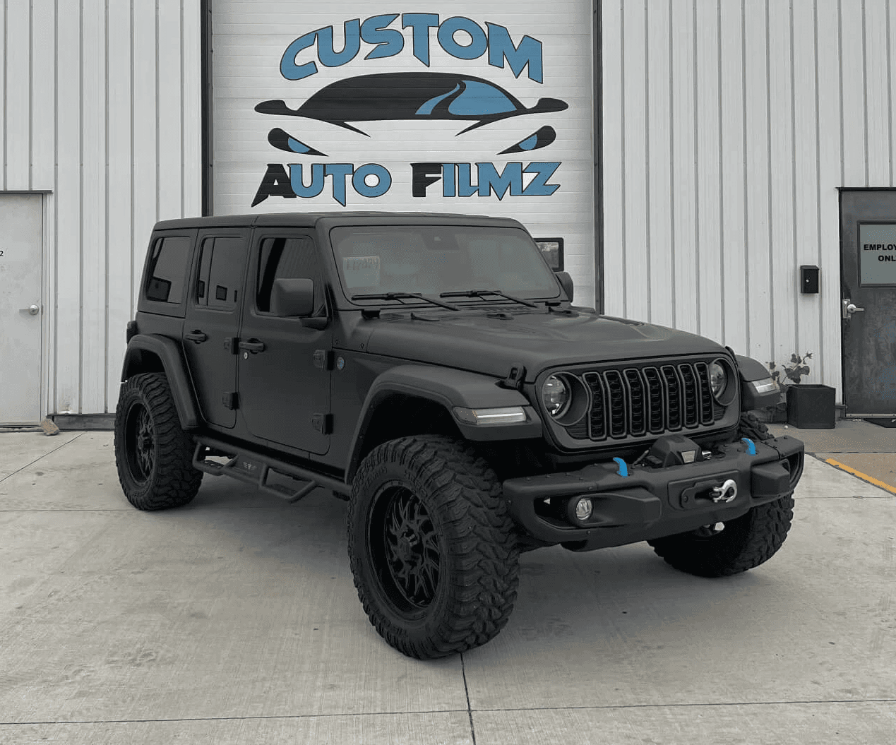 A matte black Jeep Wrangler Rubicon is parked on a concrete driveway in front of a white industrial building.