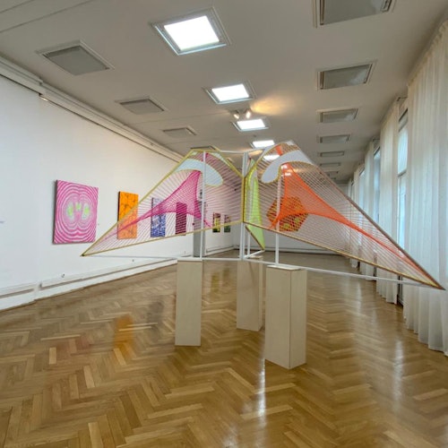 白い壁にカラフルな幾何学的インスタレーションと抽象画が展示され、研ぎ澄まされた木の床が敷かれたアートギャラリー。