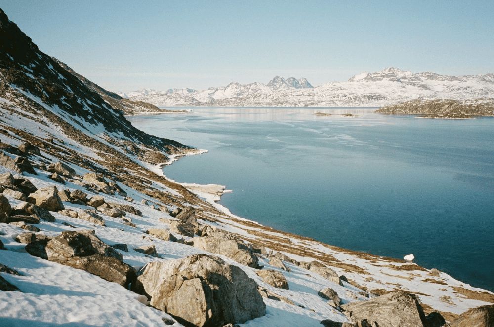 A rocky, snow-dusted coastline overlooks a calm blue fjord backed by mountains