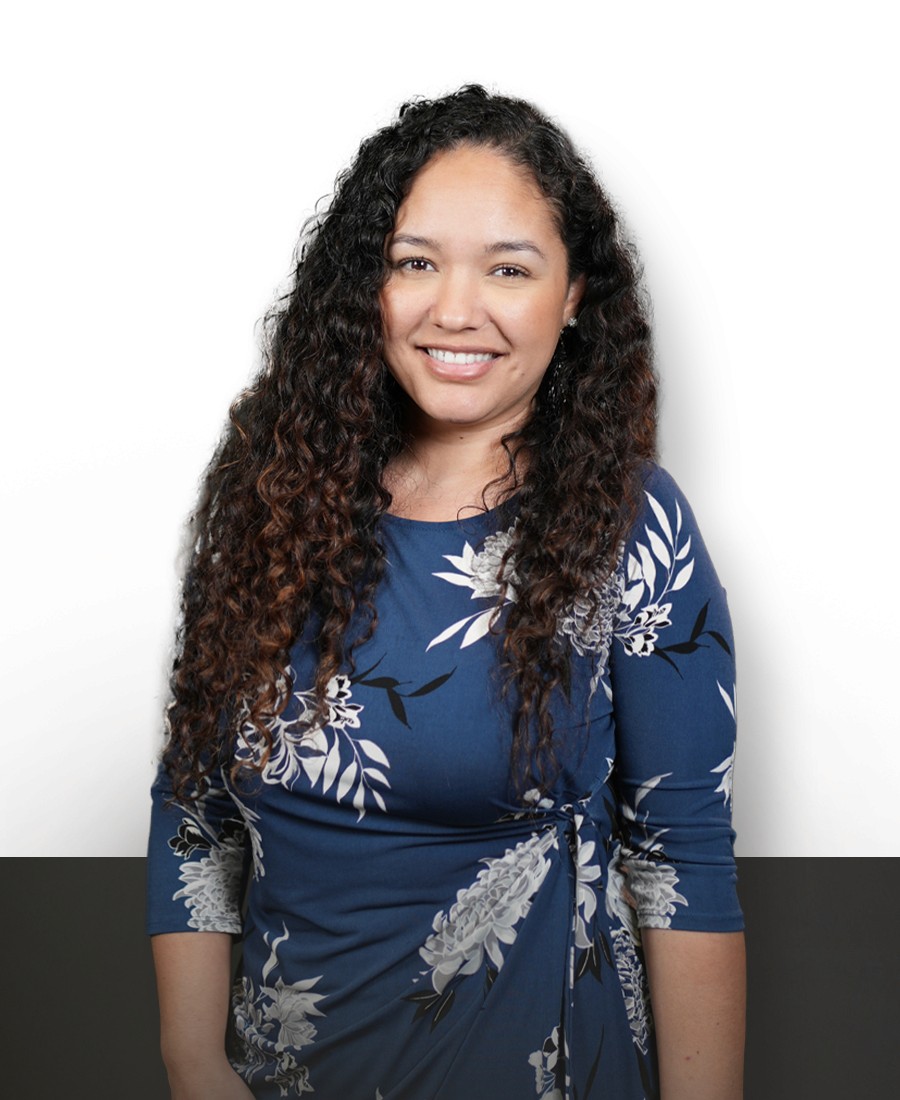 corporate headshot female with light brown skin and long curly textured dark brown hair wearing a blue wrap dress with white botanical accents, with a warm smile. 