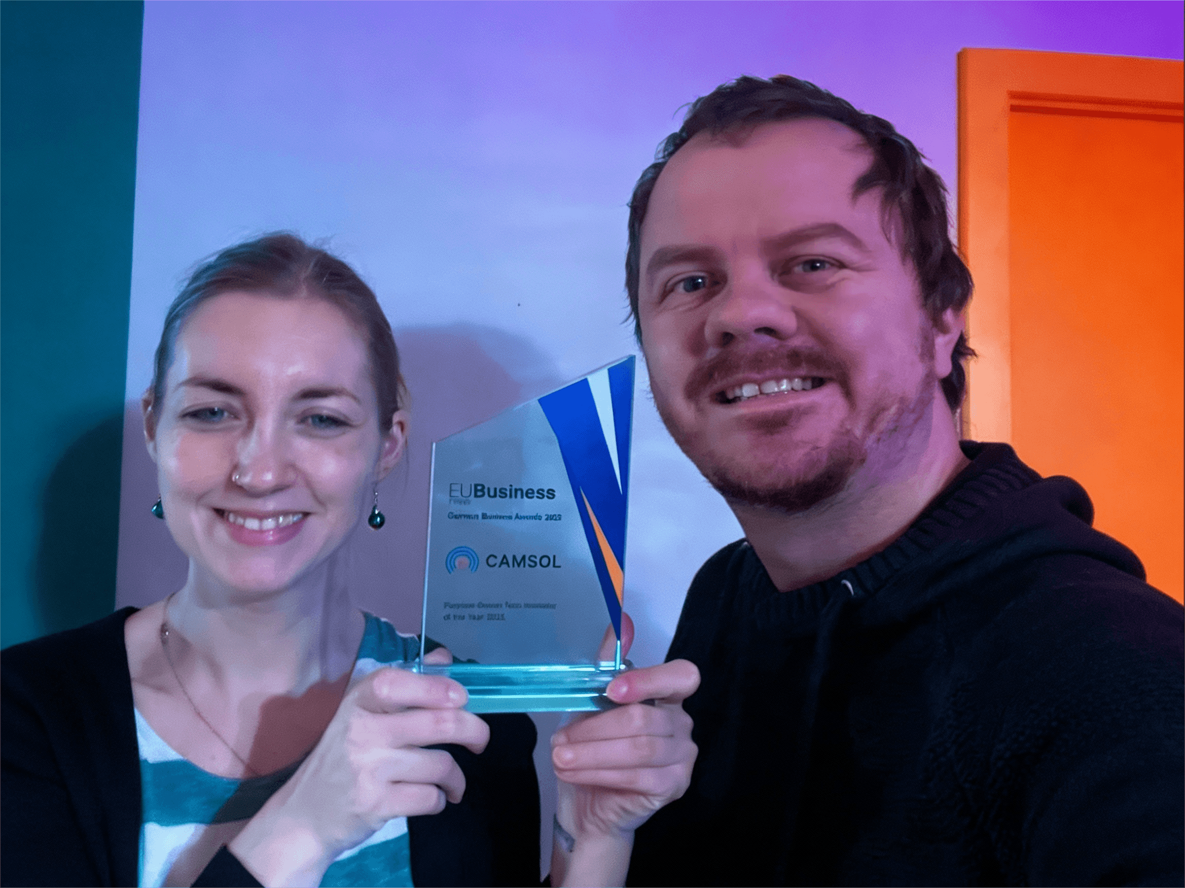 Two people smiling and holding a glass award together. The award displays the EU Business News German Business Awards 2023 recognizing CAMSOL. The background is softly lit in blue and purple tones.