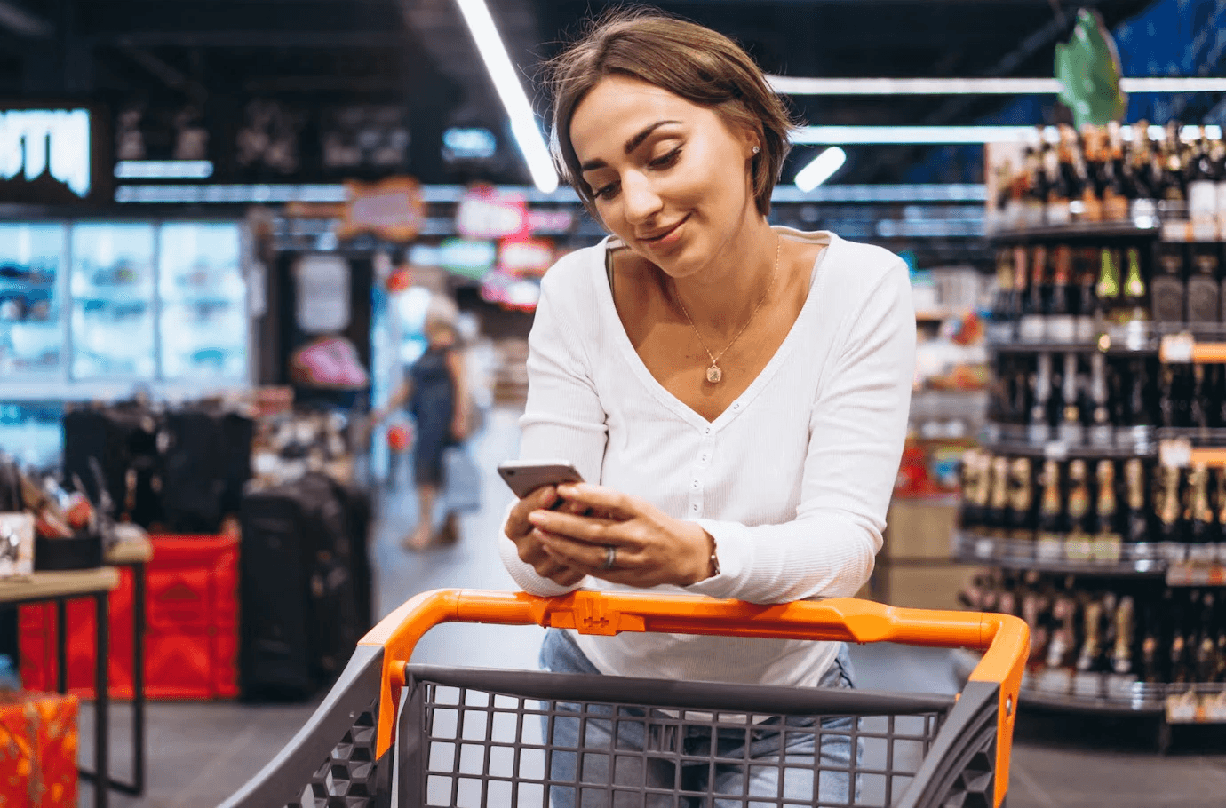 Mulher no mercado andando com o carrinho e olhando no celular