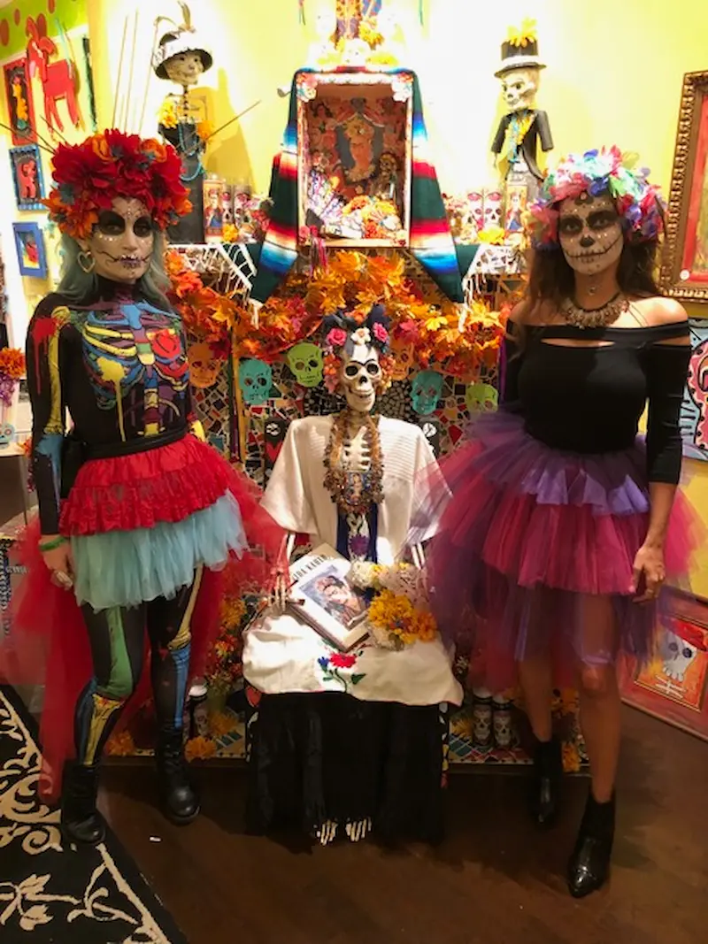 Two people dressed in vibrant, traditional Dia de los Muertos attire, complete with colorful face paint and floral headpieces, stand beside a beautifully decorated altar adorned with skull motifs and autumn leaves at the Village of the Arts in Bradenton, FL.