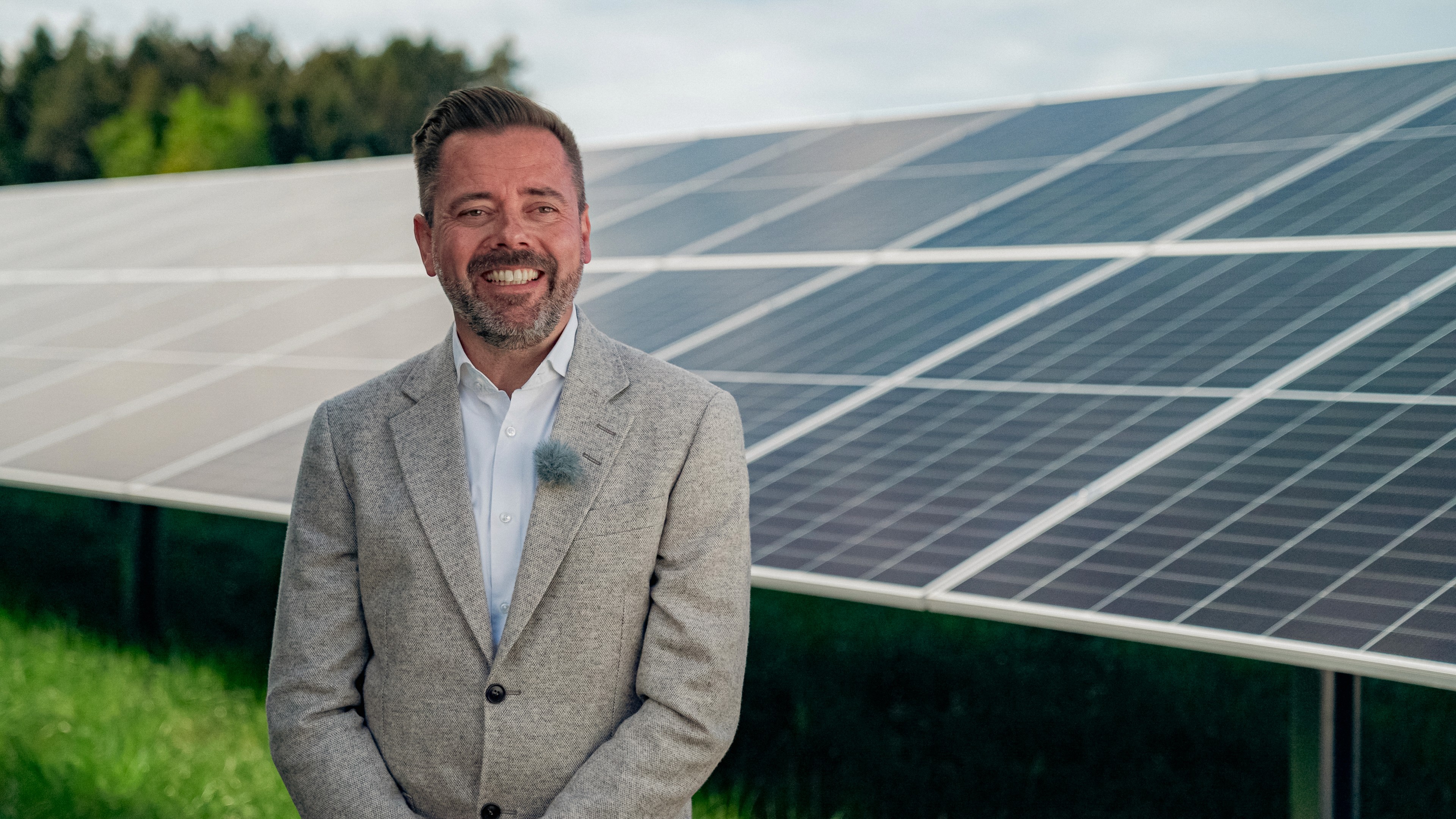 Man with brown hair and beard, wearing a beige suite, standing in front of solar panels and smiling.