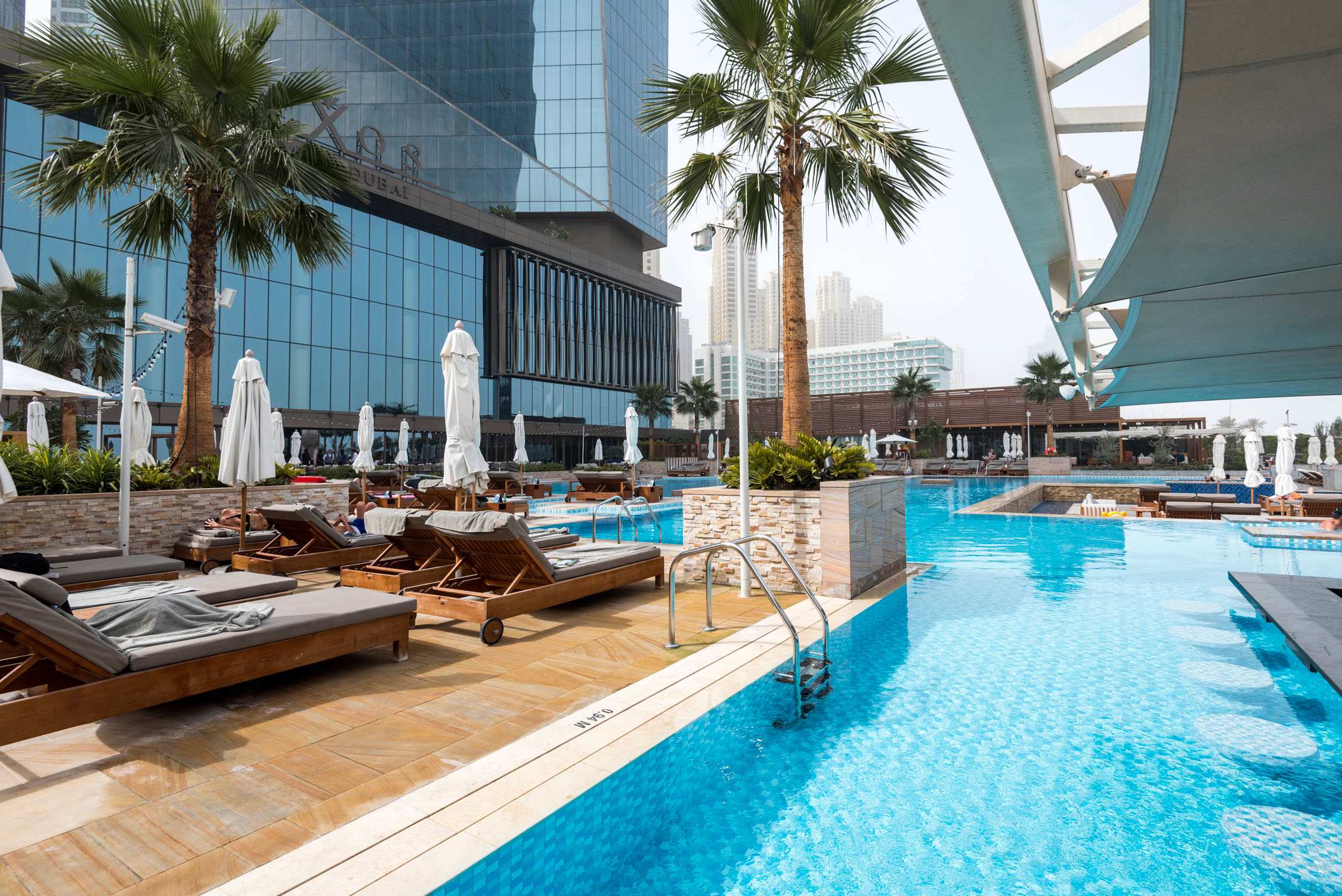 Sun loungers and white umbrellas line a turquoise swimming pool at an outdoor resort with palm trees and city buildings.