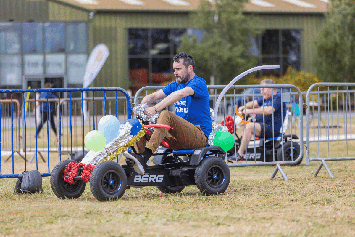 Outdoor team-building event with participants wearing matching blue shirts, taking part in inflatable obstacle courses and games, smiling and cheering, with the words 'ENTER A TEAM' in large bold white and green text across the image.
