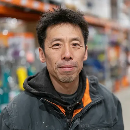 A man with short dark hair and a faint smile stands in a warehouse aisle wearing a black jacket. The background has shelves with colorful items.