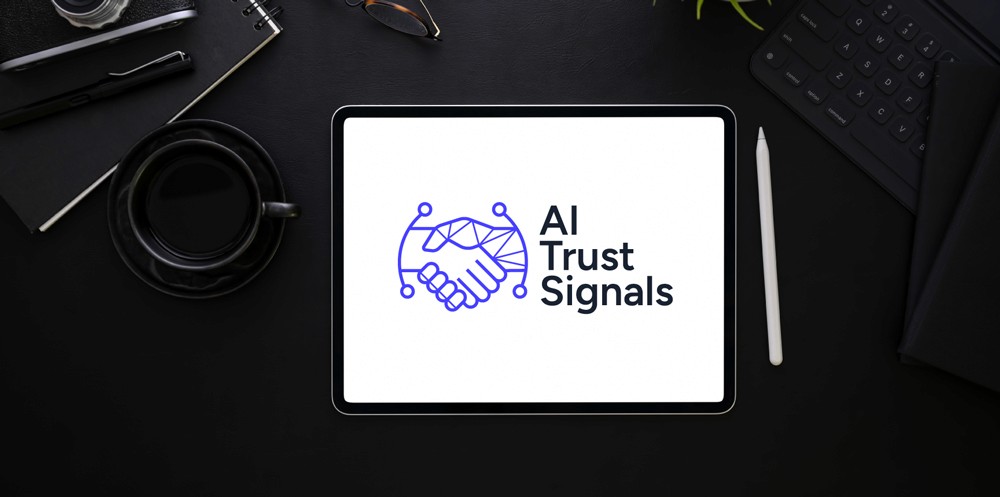 AI Trust Signals logo, featuring a digital handshake icon, displayed on an iPad screen surrounded by professional dark desk accessories, including a coffee cup, keyboard, and glasses.