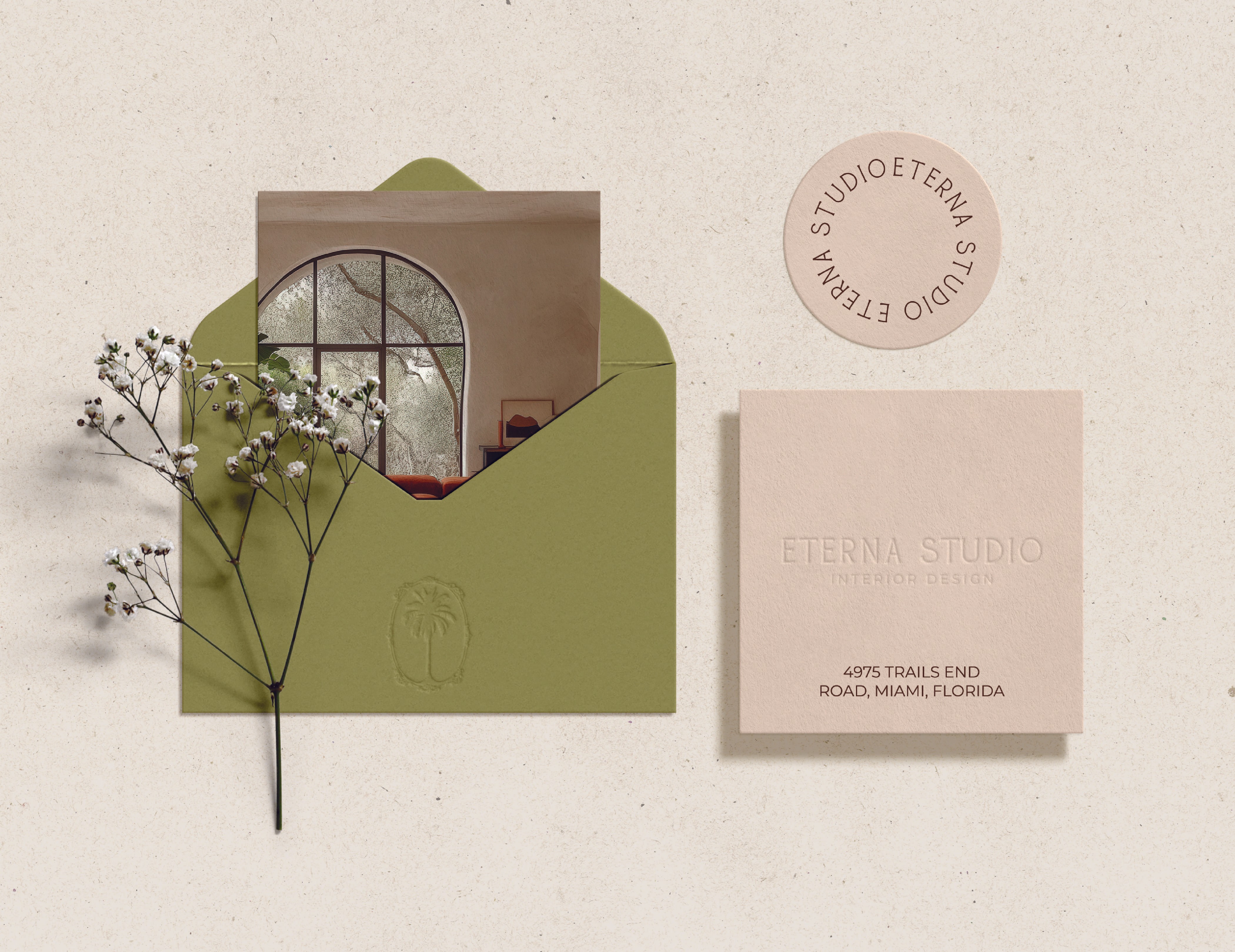 A green envelope featuring a photograph of an elegantly designed interior with a large arched window, displaying a cozy seating area. Adjacent to the envelope is a round logo stamp reading ETERNAL STUDIO and a business card with embossed text that includes ETERNAL STUDIO INTERIOR DESIGN along with an address in Miami, Florida. A delicate white flower stem leans beside the envelope, set against a textured light background.