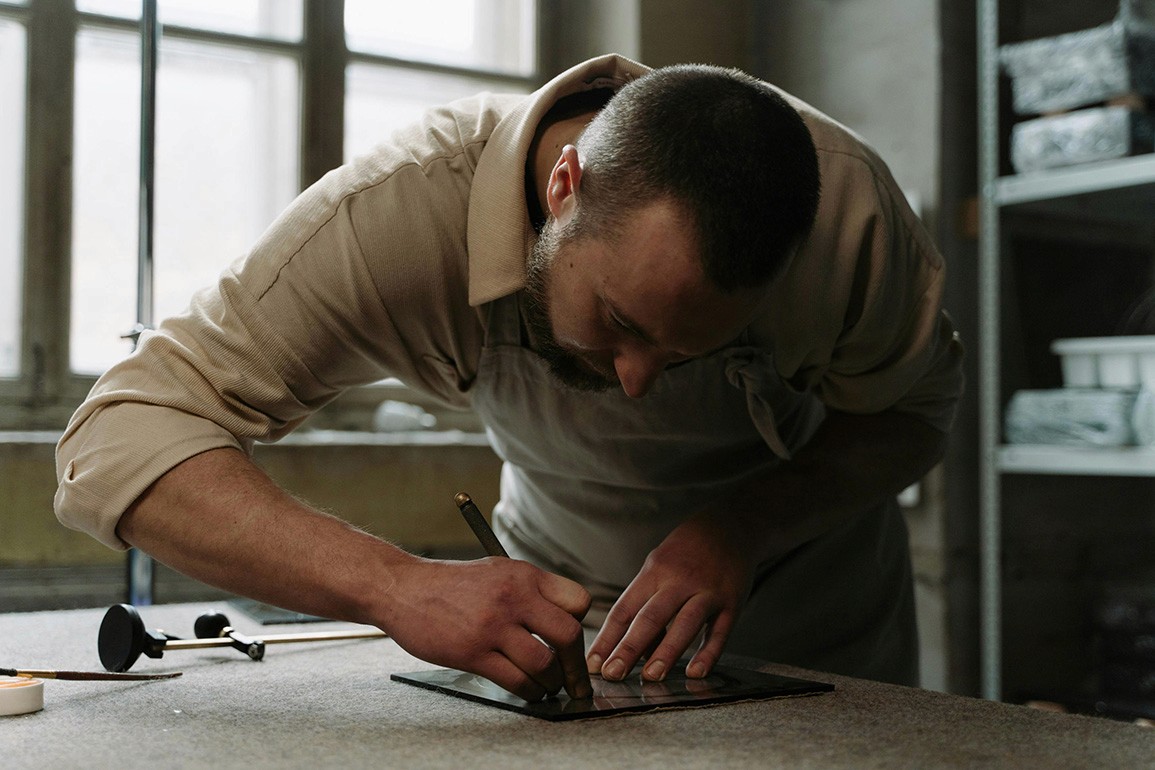 A craftsperson, wearing an apron, focuses on cutting a piece of glass with a tool on a table.