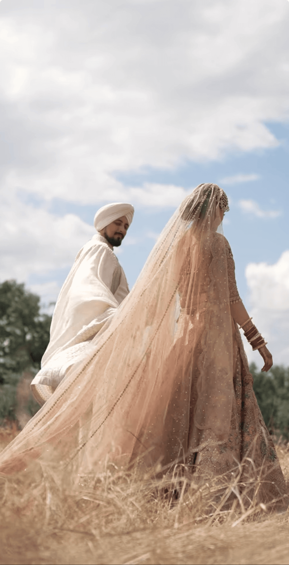 Punjabi groom in a white traditional wedding outfit and turban walking with the bride in a pink sari and veil through a sunny field with a forest backdrop.