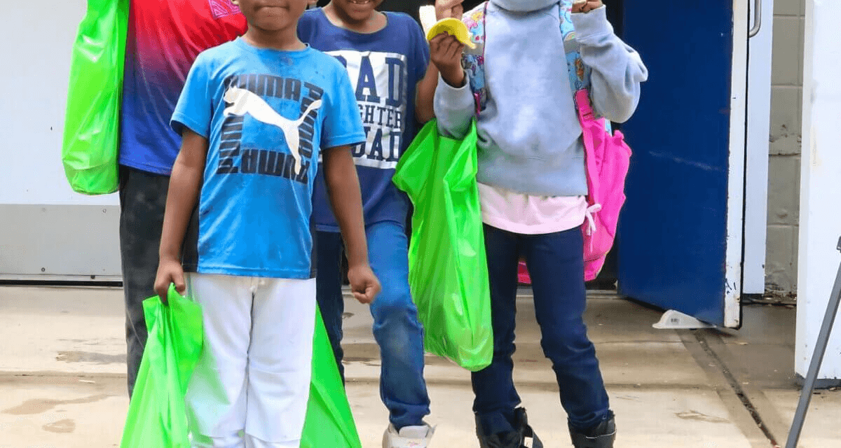 Two children walk down a sidewalk with backpacks and green food bags, participating in a local meal distribution.