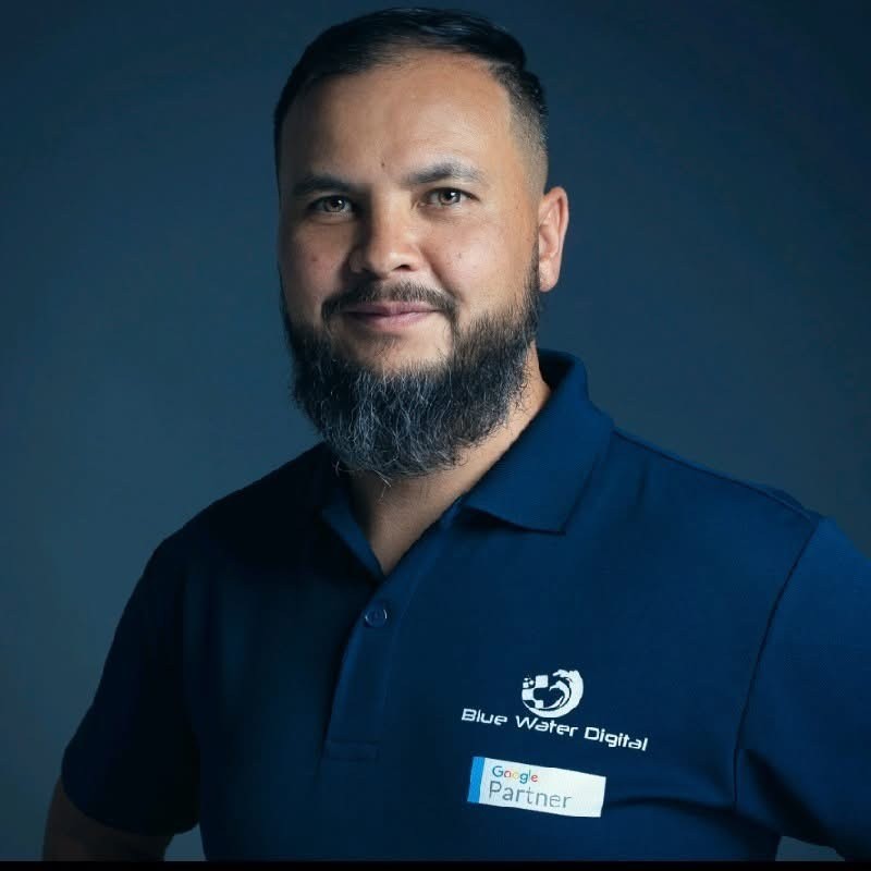 A man in a blue bomber jacket stands against a dark background. The lighting is dramatic, highlighting his focused expression. His pose is confident.