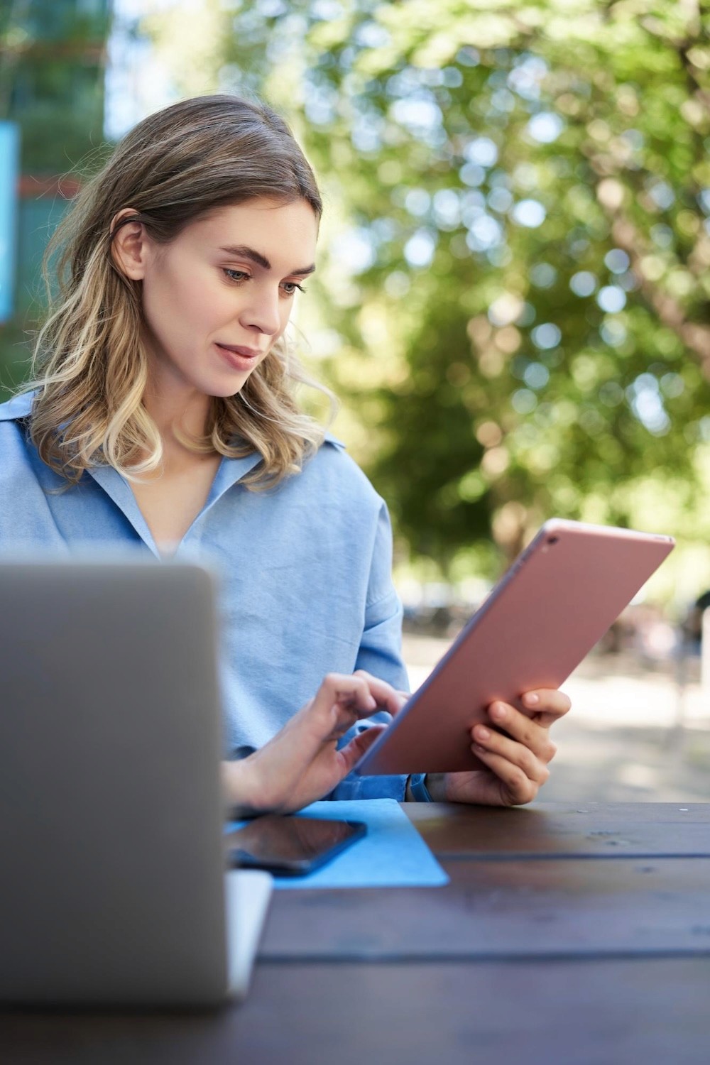 Person mit blauem Hemd sitzt draußen an einem Holztisch und hält ein rosafarbenes Tablet in der Hand