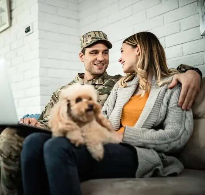 A military veteran sits on the couch with his partner and their small dog, smiling and relaxed at home. The image reflects comfort, family life, and the benefits of VA home loans offered by Chris Lewis Home Loans.