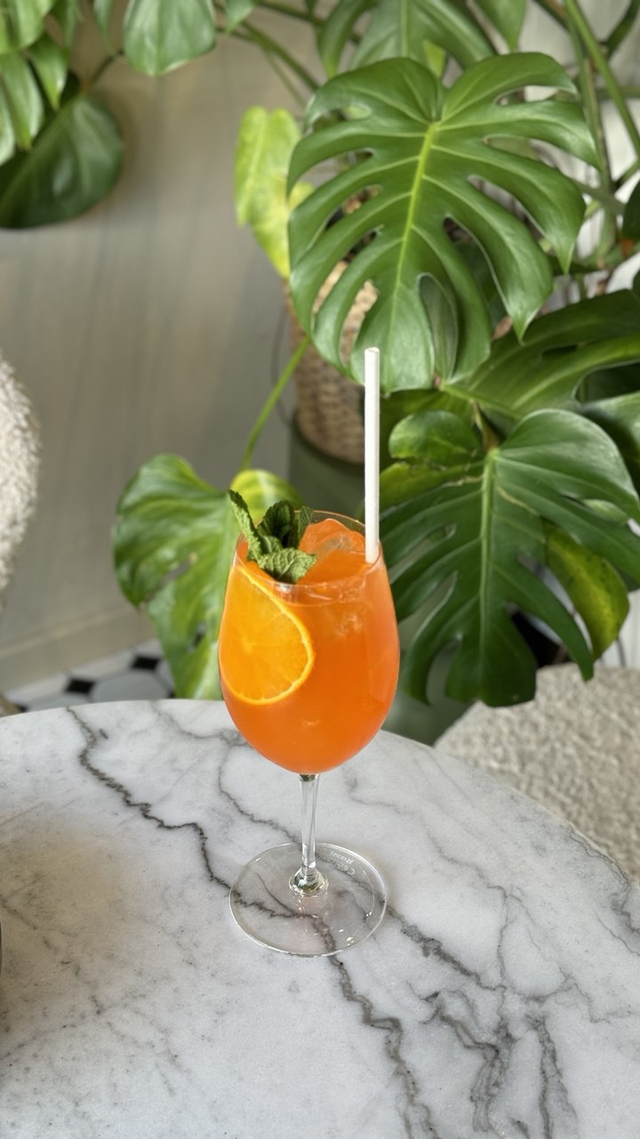 A refreshing orange cocktail garnished with a slice of citrus and fresh mint is served in a tall glass on a marble table, with vibrant green monstera leaves and a cozy interior backdrop creating a tropical ambiance.