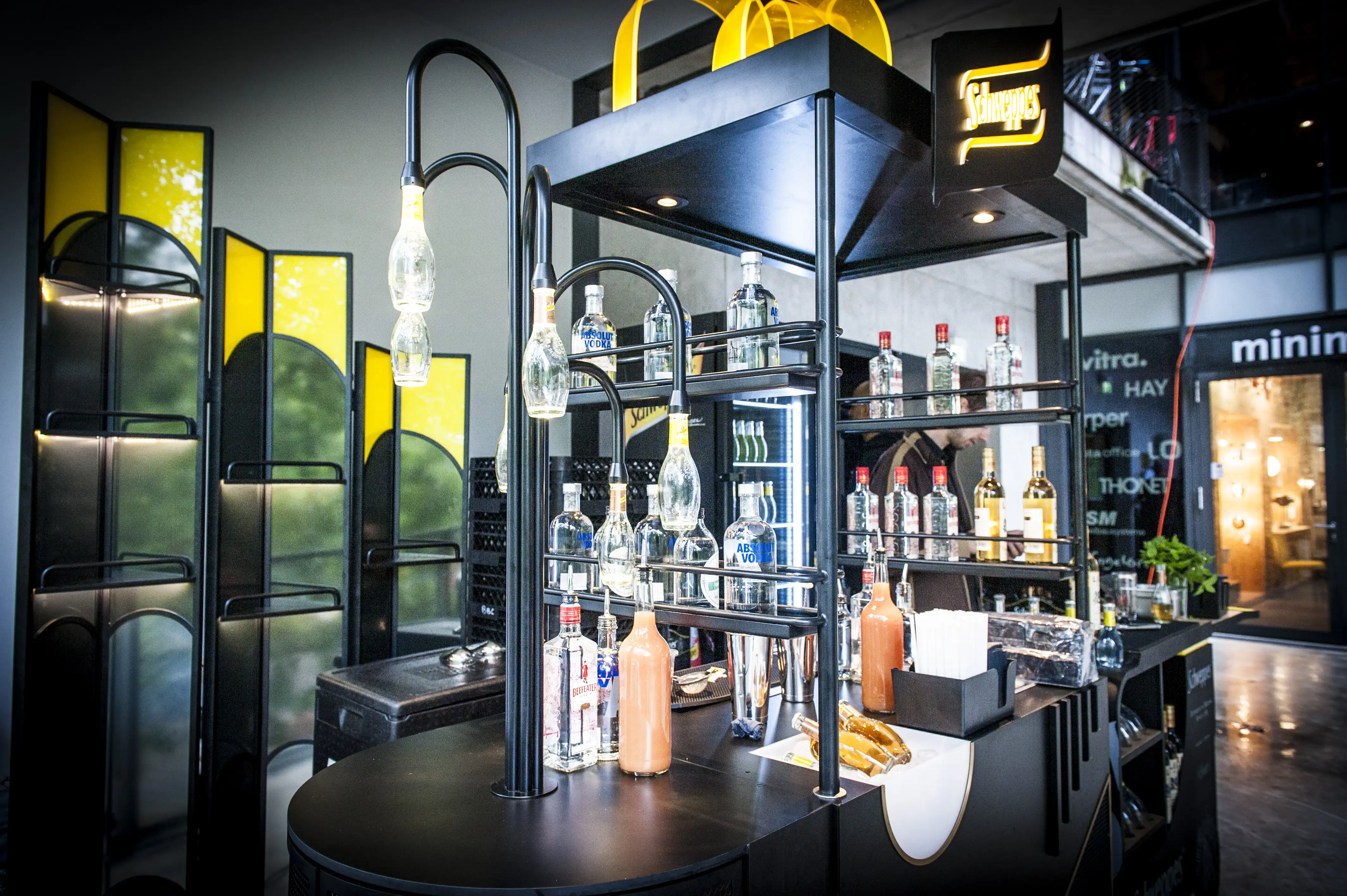 Black Schweppes bar at the STUDIOLO event with illuminated logo, bottle lamps, spirits on shelves and Art Deco folding screens with yellow accents.
