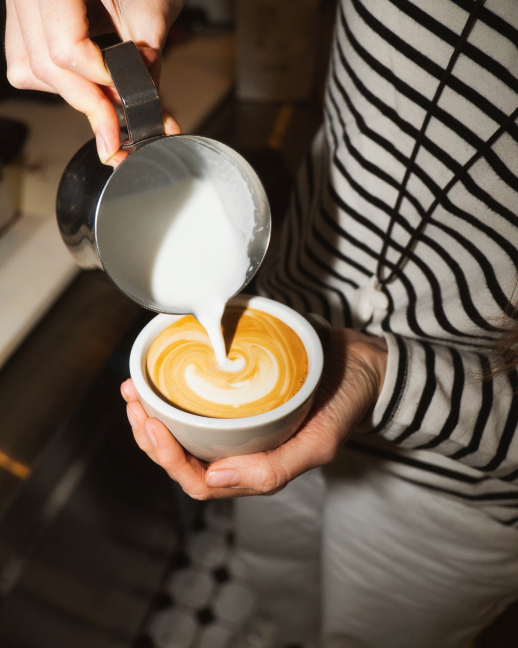 A person in a striped shirt skillfully pours creamy milk from a metal pitcher into a cup of coffee, creating intricate latte art on its surface.