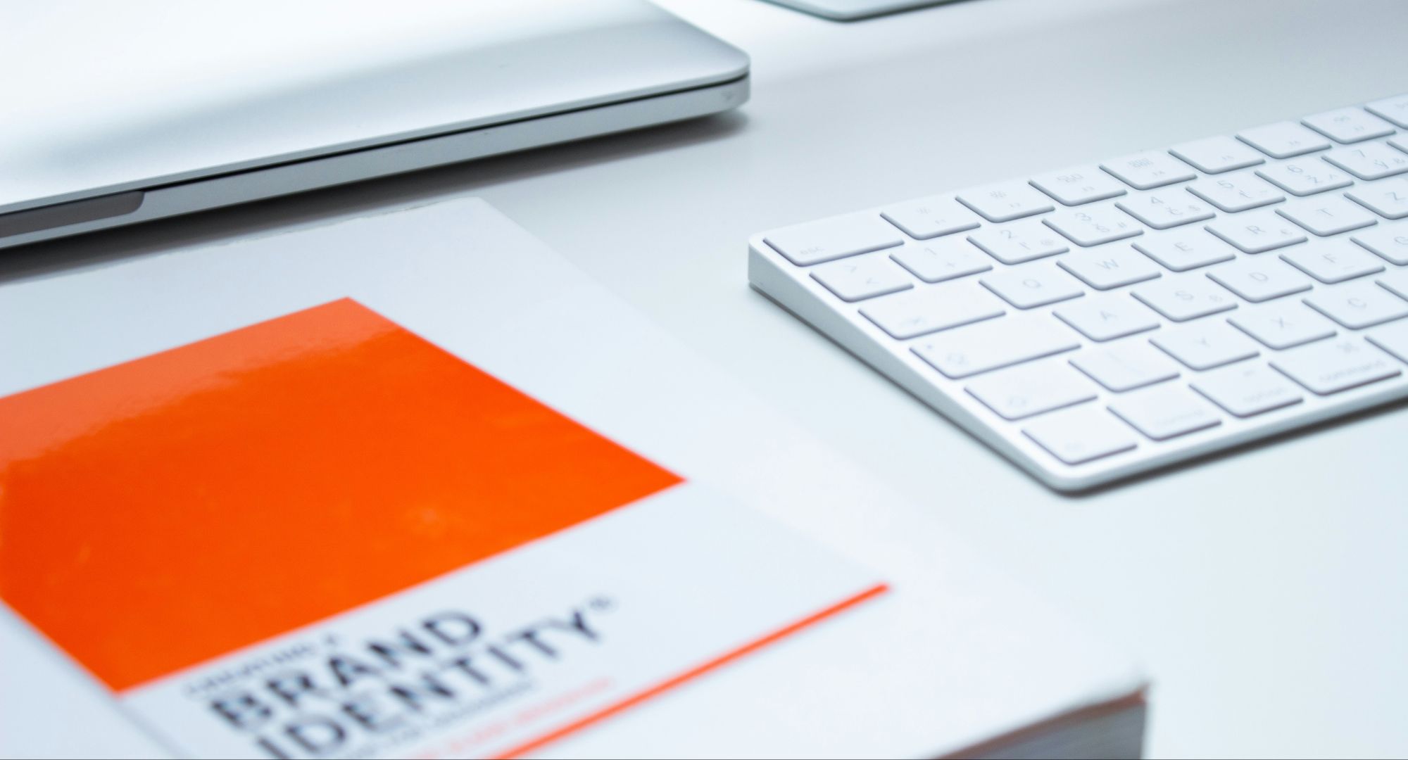 mesa branca com notebook, teclado e um caderno, brancos, com um papel laranja em destaque escrito "BRAND IDENTITY"