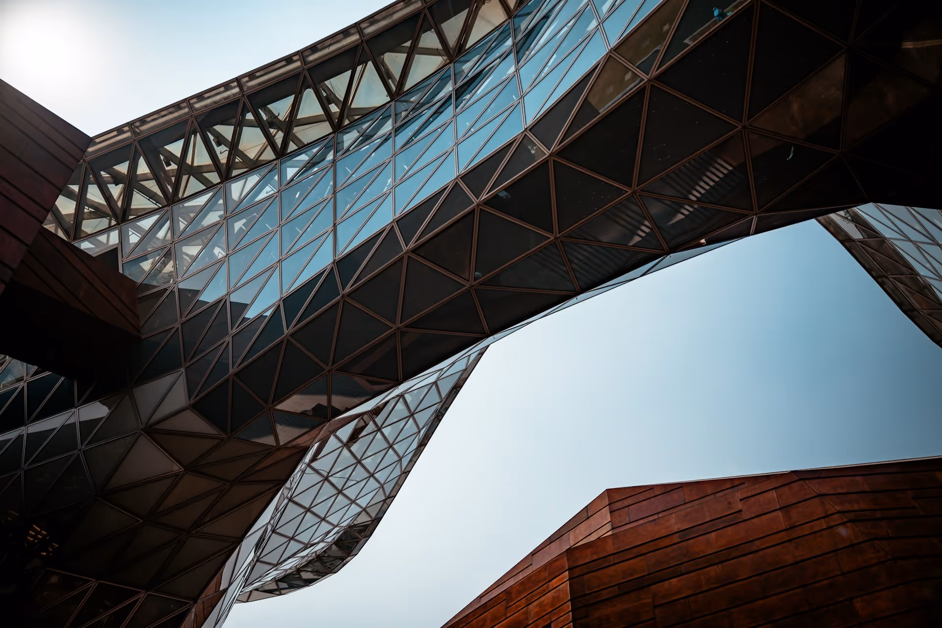 Low-angle view of a curving glass skybridge connecting modern buildings against a clear sky.