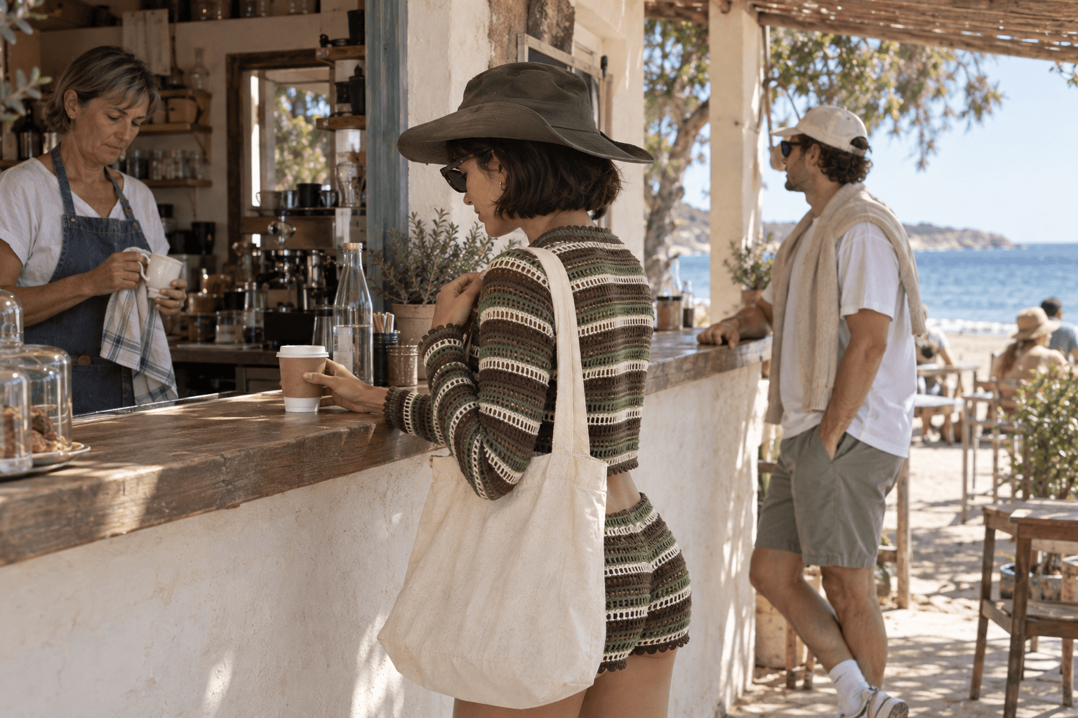 At an open-air beach coffee hatch, a woman in striped knitwear reaches for a cup on the wooden counter while holding her shoulder-bag strap; an older female barista dries a ceramic cup and her companion looks toward the sea.