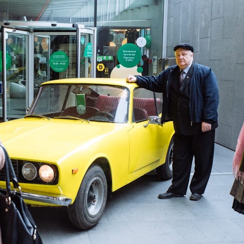 Egy idősebb férfi áll egy sárga, régi autó mellett egy üveg bejárat előtt.