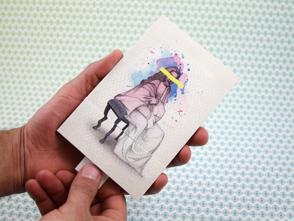 Two hands hold a small rectangular card. The card displays a watercolor illustration of a person with long dark hair, wearing a purple robe and seated on a dark chair with legs visible. The figure has a yellow band across their forehead. The background is light blue with a faint repeating pattern. The hands holding the card are positioned against a light-colored surface with a subtle texture.