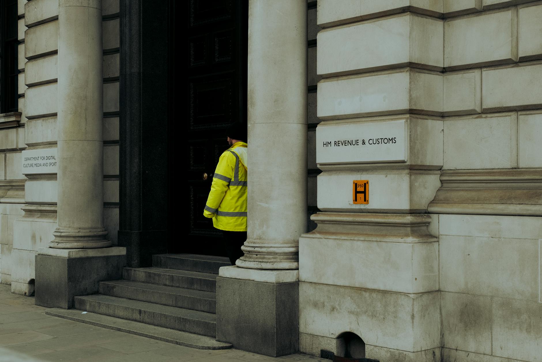 Worker in a yellow high-visibility jacket entering the stone-columned entrance of the HM Revenue & Customs building
