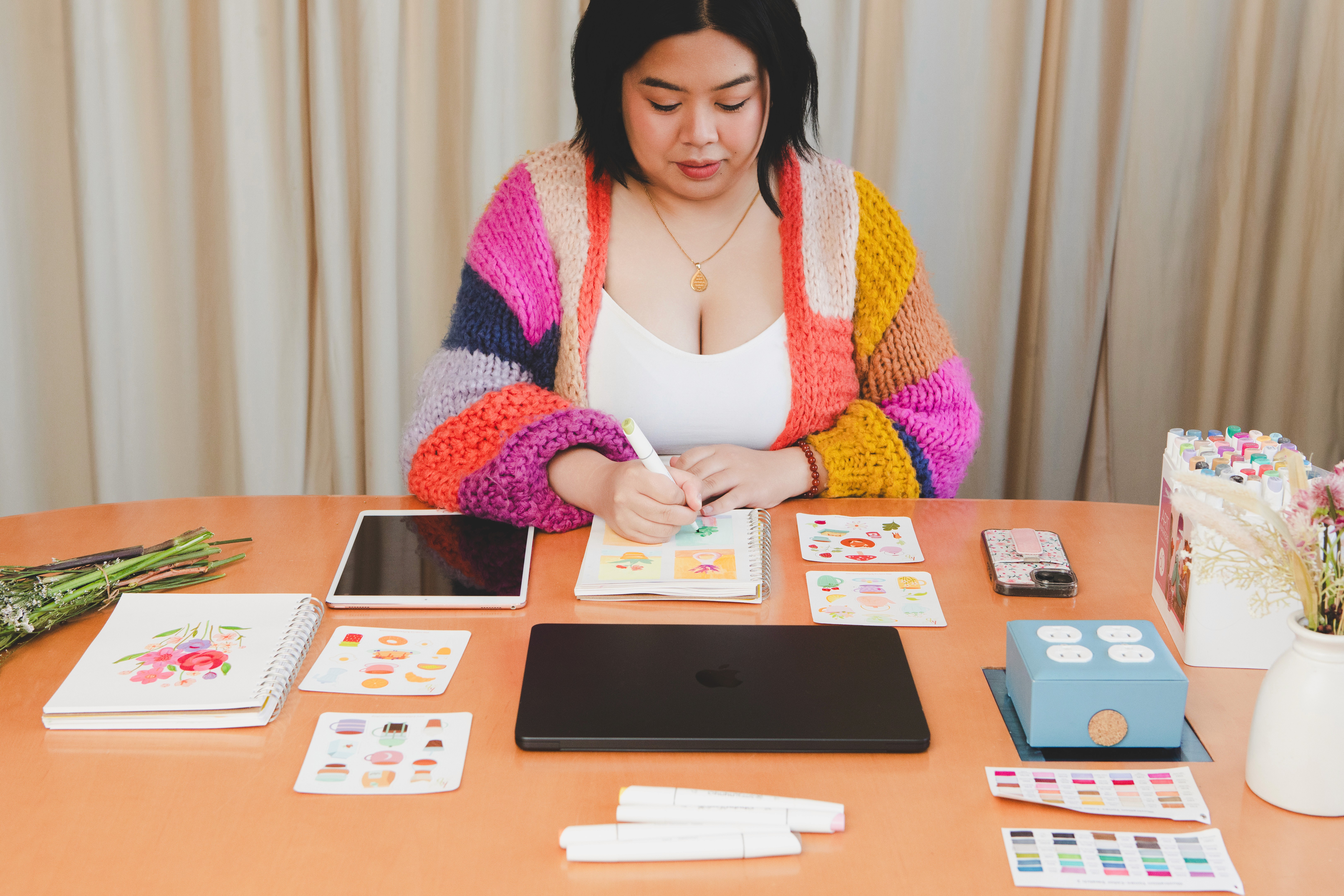 Designer wearing colourful cardigan as she colours with markers, stickers, and laptop around
