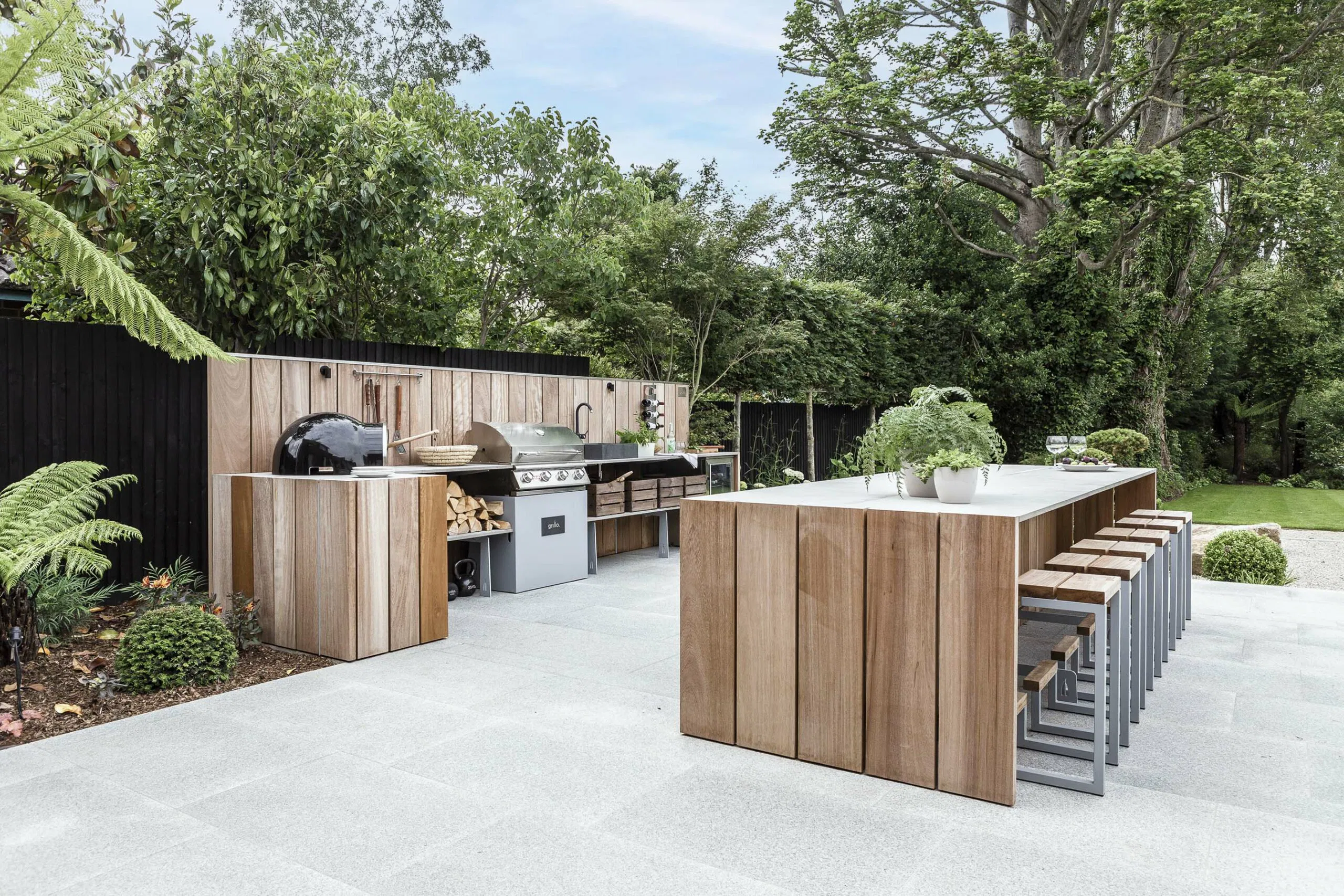 Outdoor kitchen with wooden structures, a grill, and a countertop in a lush green garden setting.