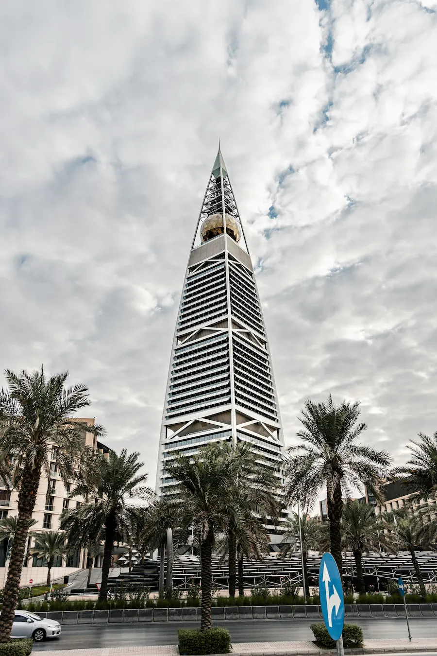 Al Faisaliah Tower in Riyadh, Saudi Arabia, rising above palm trees under a cloudy sky.