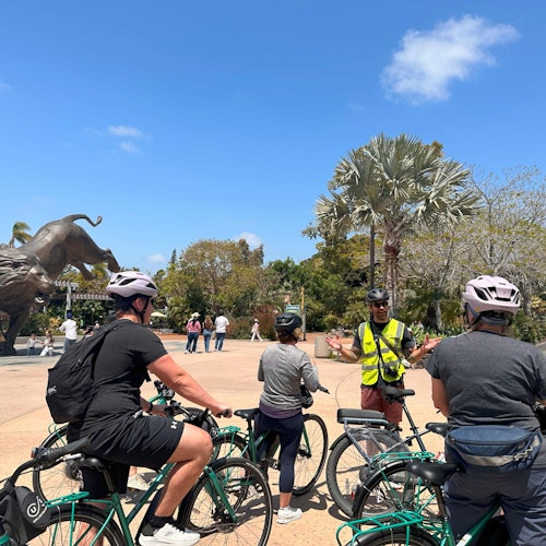 Egy csoport kerékpáros megáll, hogy meghallgassa a sárga mellényes idegenvezetőt. A háttérben egy nagy állatszobor és fák láthatók.