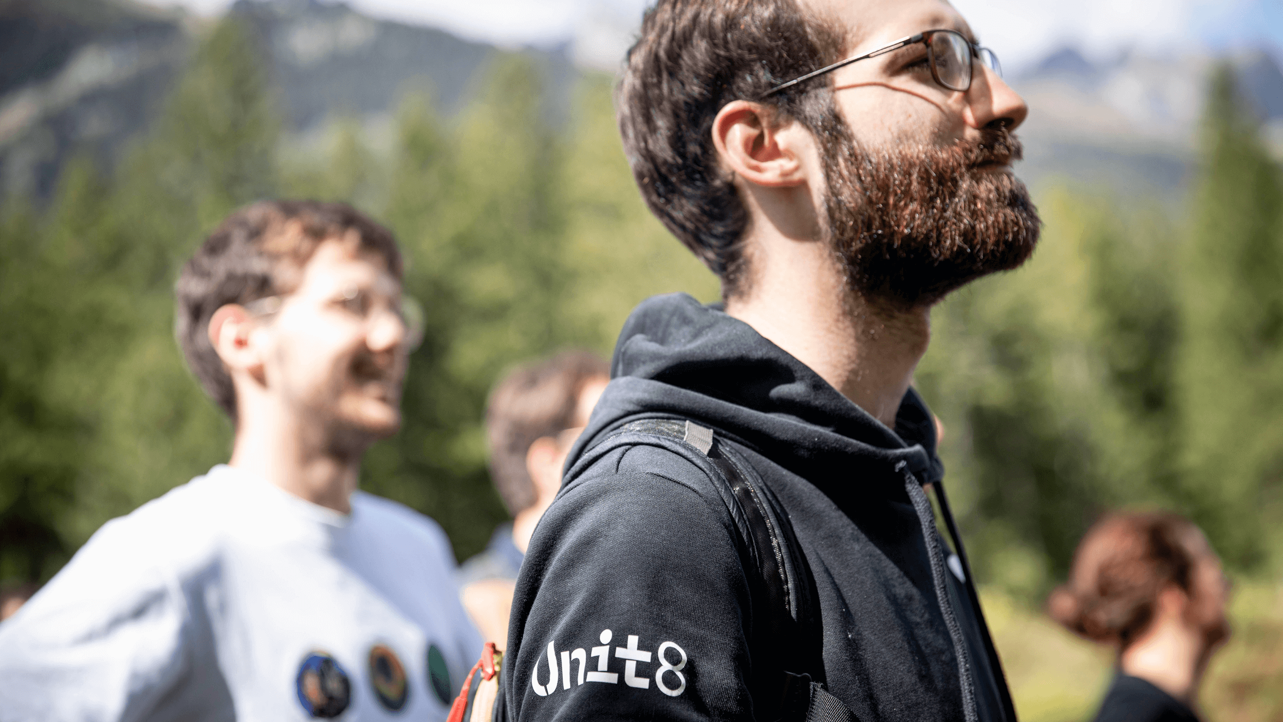 Mężczyzna z brodą w czarnej bluzie z logo Unit8, w tle koledzy i górski krajobraz
