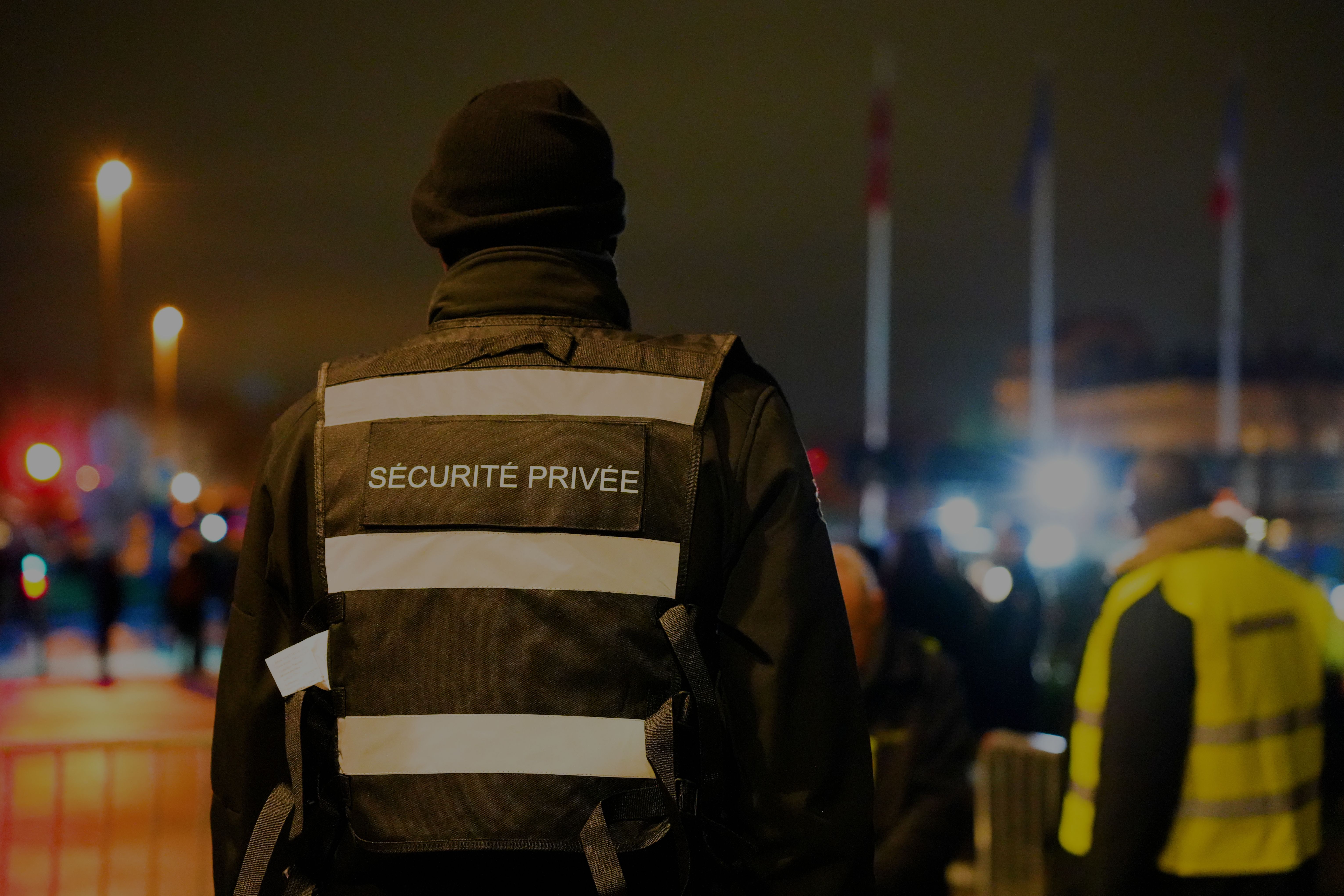 Agent de sécurité de dos avec un oddard Sécurité Privée en fond un autre agent desécurité avec un gilet jaune et la rue floue
