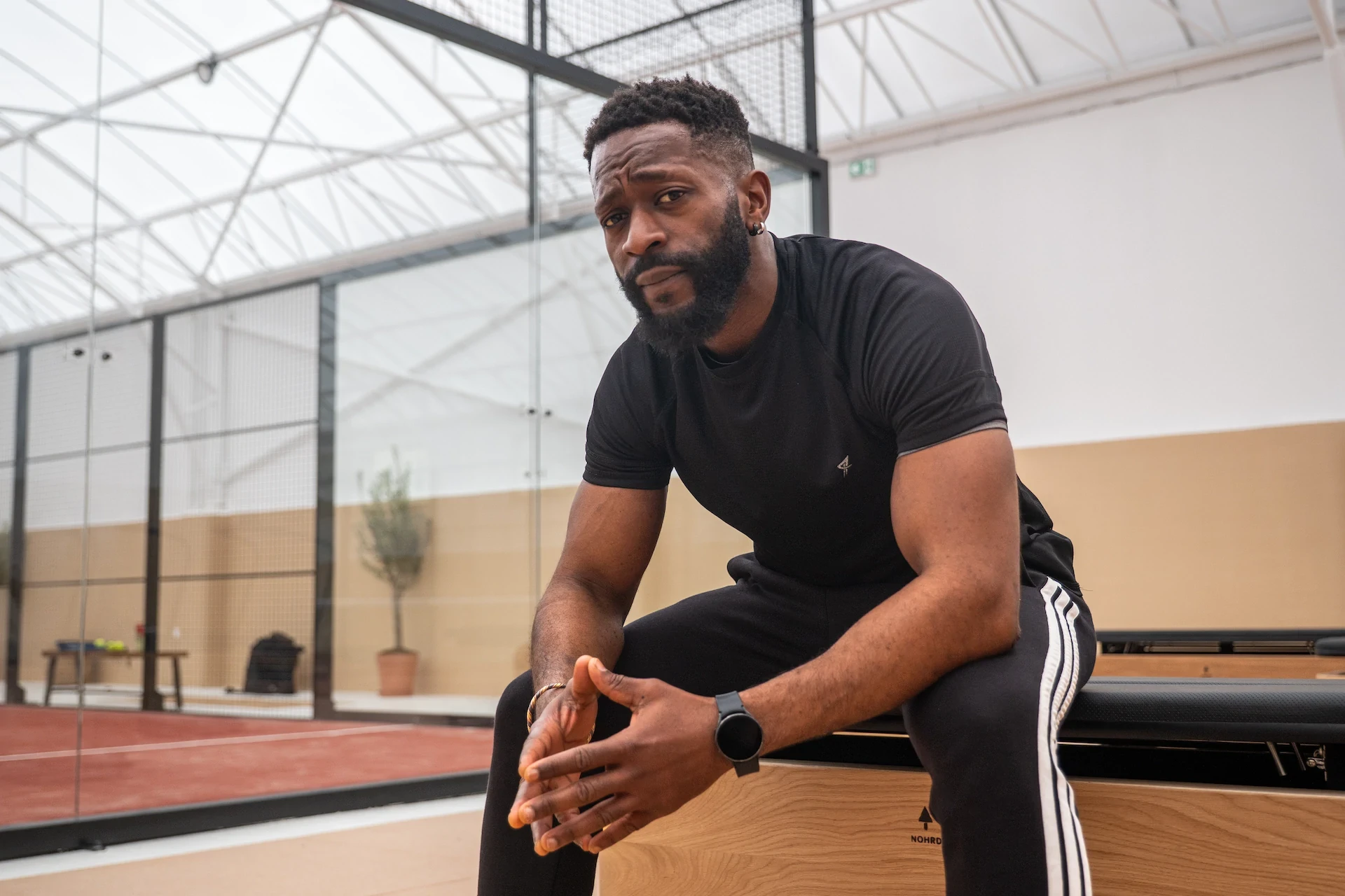 Homme noir assis, regard sérieux. Tenue de sport noire. Fond de terrain de sport.