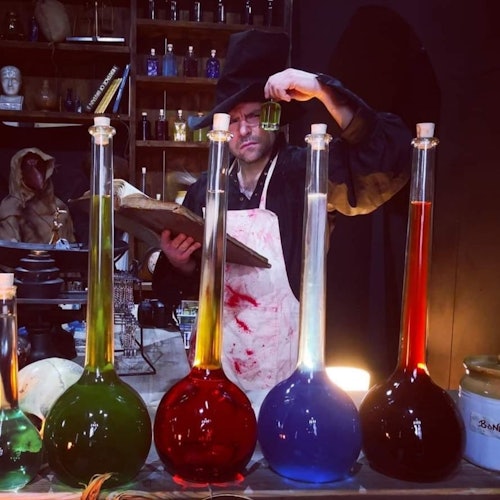 一位戴着帽子和围裙的人在实验室中检查一个绿色的小瓶，桌上摆放着大瓶的彩色液体，背后是架子上的瓶瓶罐罐。