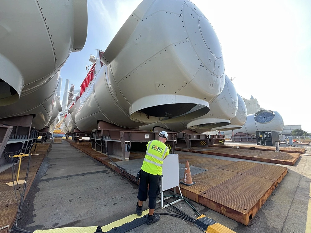 Large wind turbine components being handled at a port, with a worker in high-visibility clothing in the foreground.