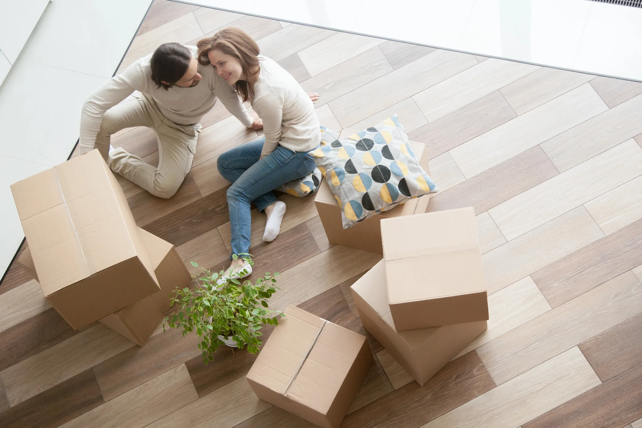 White couple sitting on a floor of a house, wife leaning on the husband's shoulder, unopened boxes beside, a plant in front of them