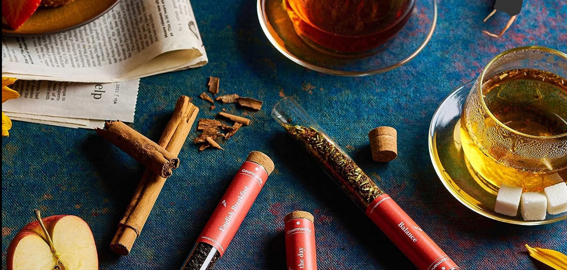 A vibrant display of assorted tea leaves in glass tubes surrounded by cinnamon sticks, sliced apples, sugar cubes, and a glass cup of brewed tea on a rich blue tablecloth, accompanied by a folded newspaper.