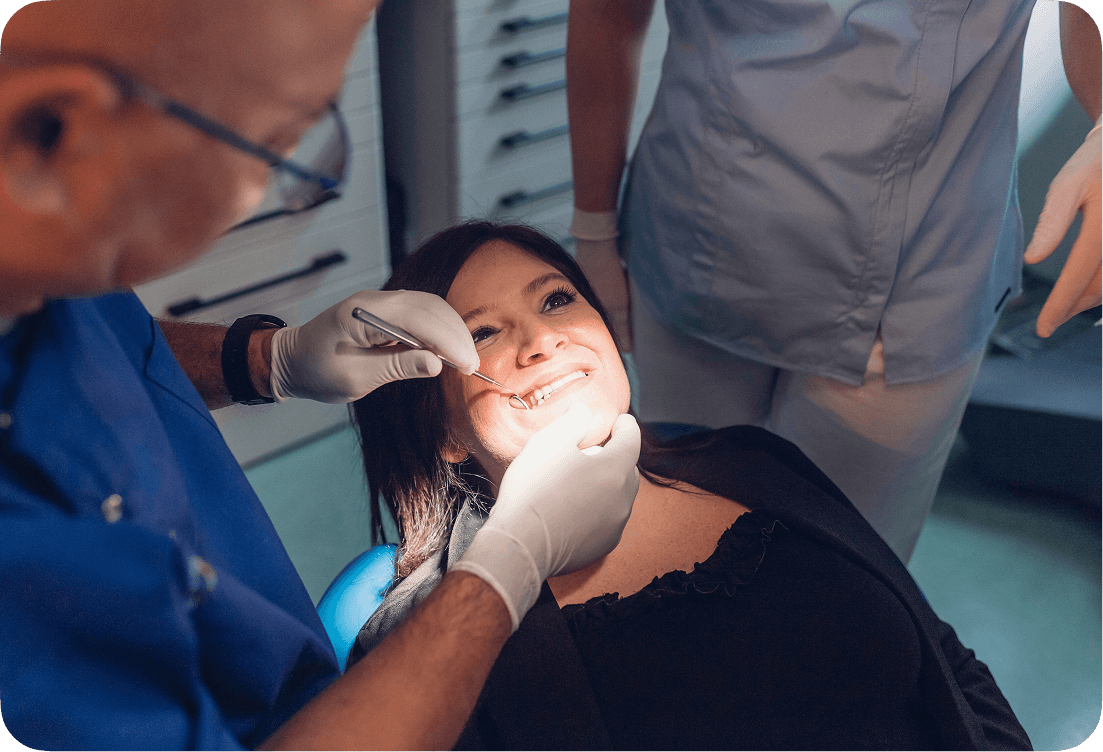 A dentist in blue scrubs uses dental tools to examine a smiling patient's teeth during a check-up, with an assistant nearby in a well-lit dental office.