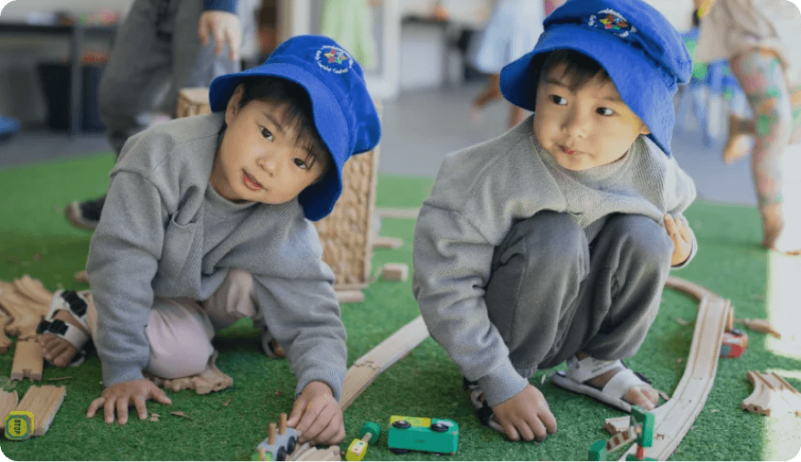 Two children wearing blue hats building wooden train tracks during indoor play at Kaleidoscope Kids Goodna childcare centre.