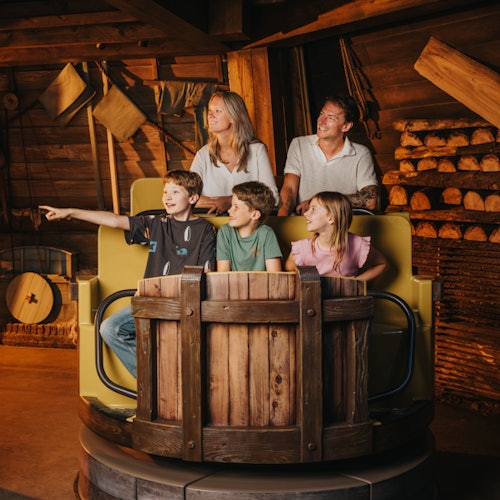 Una familia de cinco personas está sentada en un vehículo de una atracción de parque temático, sonriendo y mirando algo fuera de escena; el entorno se asemeja a una cabaña de madera.