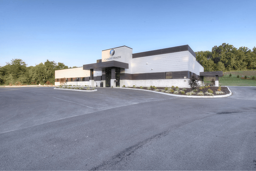 Modern building with a symmetrical design, surrounded by a parking area and greenery under a clear blue sky.