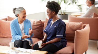 Older woman and caregiver sitting on a sofa, chatting and holding cups in a comfortable living room.