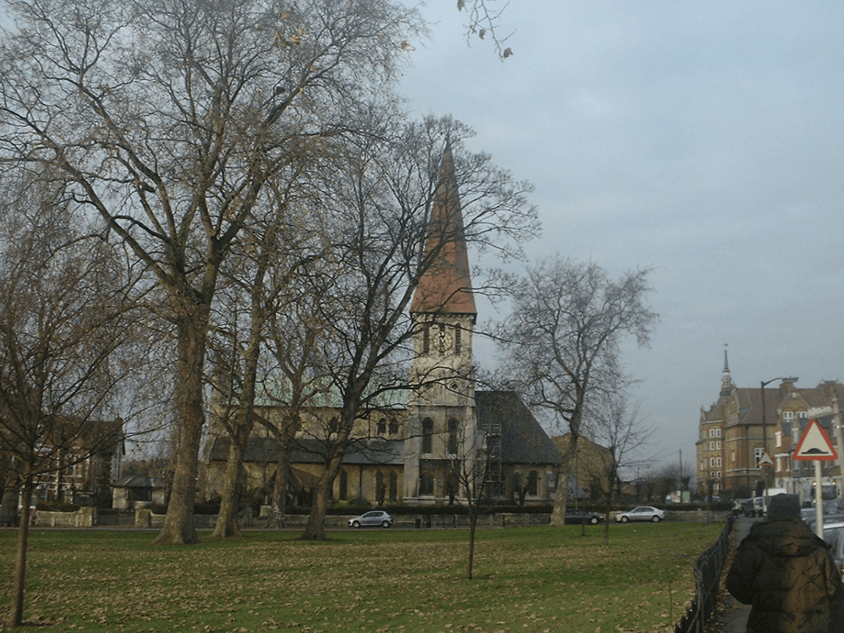 A smiling man with curly hair is sitting at a desk, looking at the camera, with a laptop and a lamp in the background.