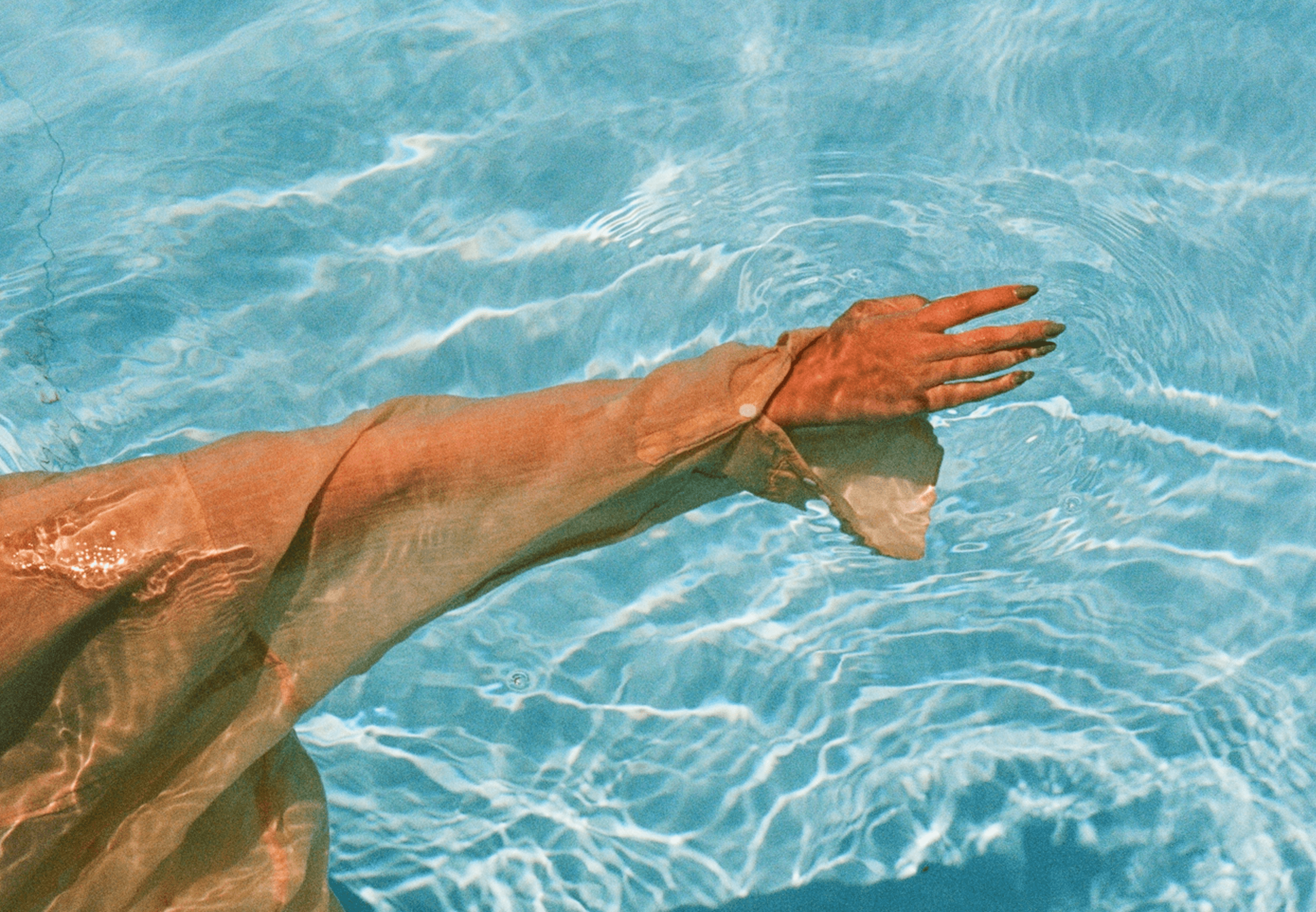 A person wearing a sheer, long-sleeved garment extends their arm into a clear blue pool, creating gentle ripples in the water with sunlight reflecting on the surface.