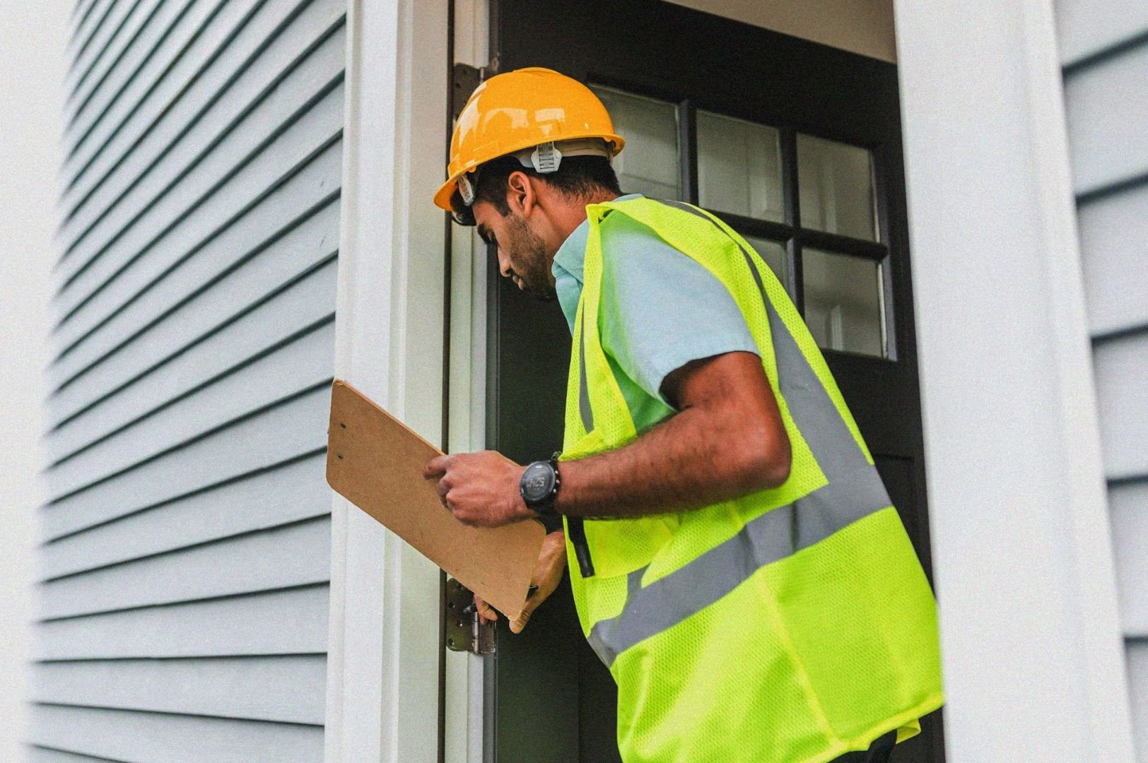 A home inspector wearing a yellow hard hat and safety vest examines a house entrance, holding a clipboard. The siding is light gray, suggesting a residential setting.
