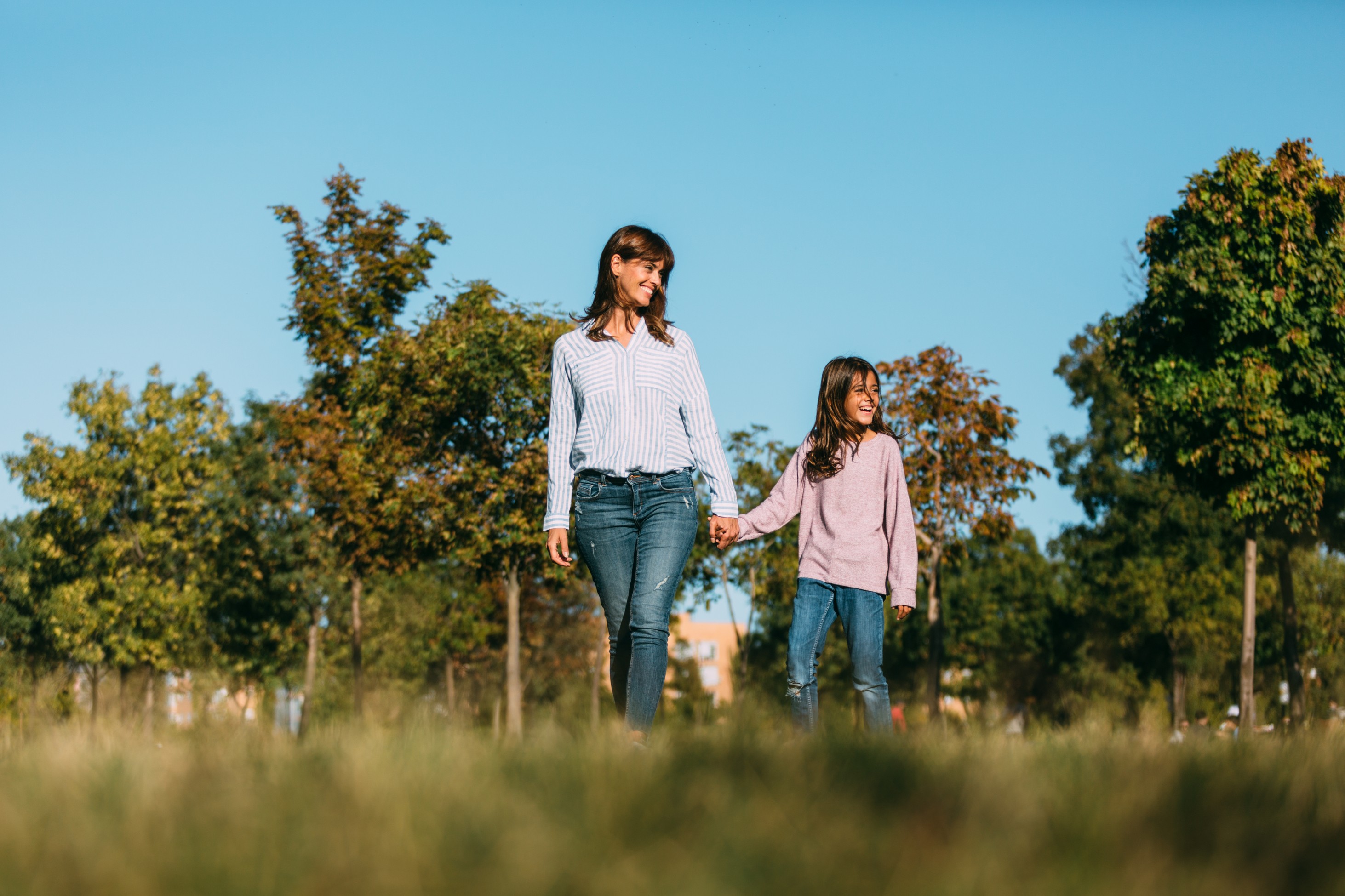 A woman and her daughter hold hands while walking through grass, with leafy trees and open sky behind them